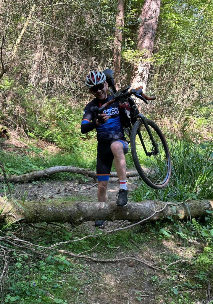London Dynamo rider climbing over a log with his bike over his shoulder on a gravel ride in Surrey